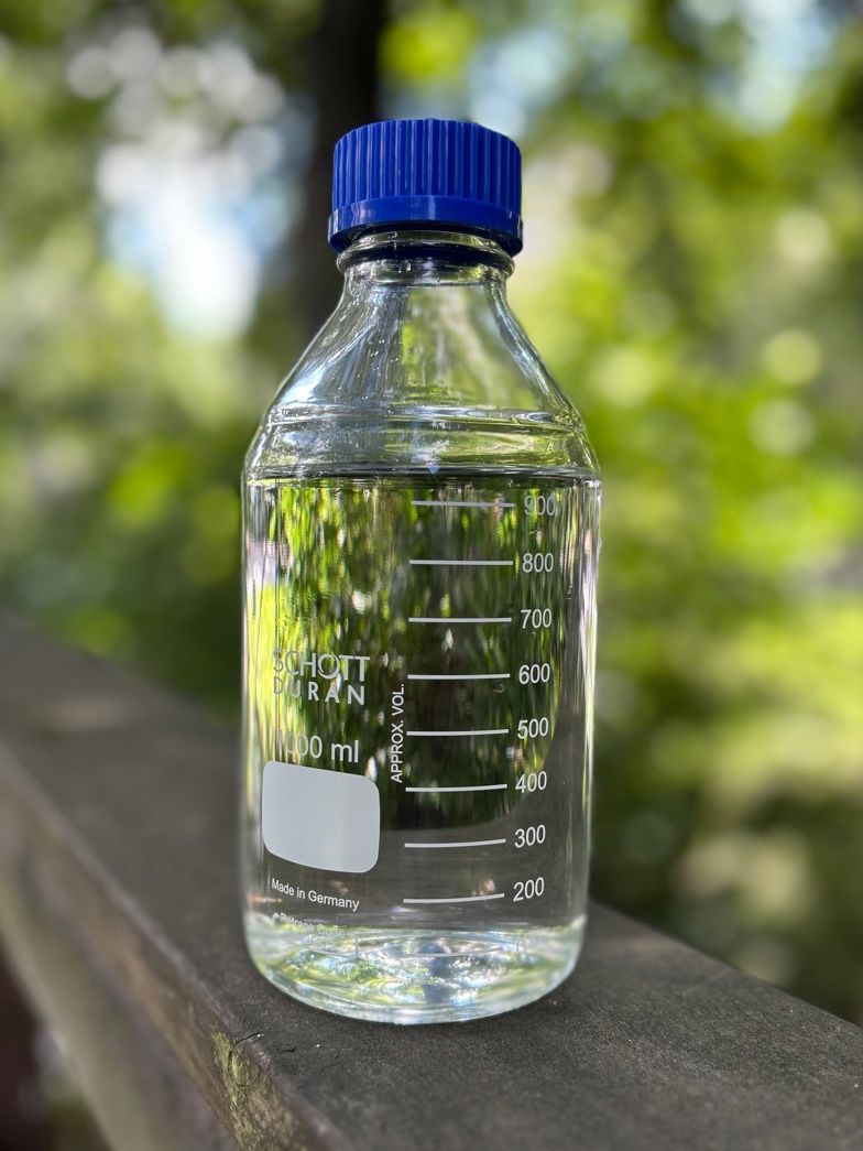 Vattenflaska av typen Schott Duran i glas med blått skruvlock. I bakgrunden syns träd och grönska. 