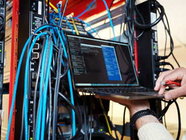 En man håller i en laptop i ett serverrum fullt med sladdar.